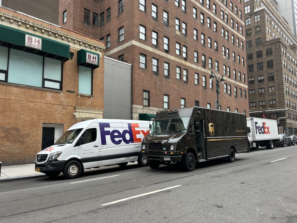 FedEx and UPS delivery trucks double-parked on a busy New York City street, highlighting last mile delivery congestion and emissions challenges addressed by Smart Package Room systems sustainable package logistics solutions.